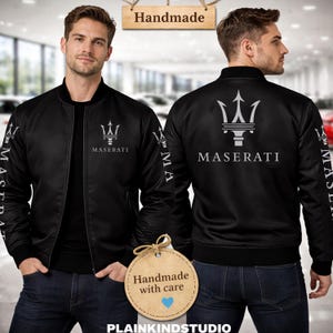 May include: A black bomber jacket featuring the silver Maserati trident logo and text on the chest and sleeves. The jacket has a ribbed collar, cuffs, and hem. A wooden sign above reads Handmade, and a circular tag below reads Handmade with care.