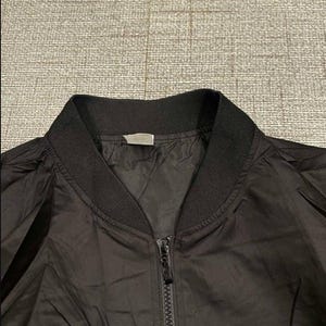 May include: A black bomber jacket with a ribbed collar and cuffs. The jacket has a full zipper closure down the front. The fabric appears to be a lightweight material, suitable for layering. The jacket is a classic style.