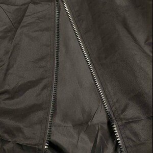 May include: Close-up of a black jacket with a zipper. The jacket appears to be made of a smooth, possibly water-resistant material. The zipper runs down the center, partially open, revealing the interior lining.
