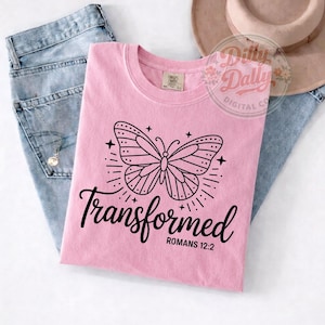 Può includere: Una maglietta rosa chiaro con una grafica a farfalla nera e la parola "Transformed" in corsivo. Il testo "Romans 12:2" è sotto la parola "Transformed". La maglietta è piegata sopra un paio di jeans blu.