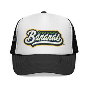 Peut inclure: Casquette de camionneur noire et blanche avec une visière incurvée. Le panneau avant est blanc et porte l'inscription "Savannah Bananas" dans une écriture stylisée, soulignée de jaune, de bleu et de vert. Le chapeau a un dos en filet noir et une visière noire.