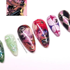 May include: A collection of artificial nails with various designs. Some nails feature comic book art of two characters embracing, while others have abstract patterns in pink, purple, and silver. One nail is green with a floral design.