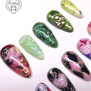 May include: A collection of custom-designed, long, almond-shaped artificial nails. The nails feature various designs, including a portrait, geometric patterns, and glitter accents. Colors include green, pink, gold, and black. The Kutty Handmade Nails logo is in the upper left corner.