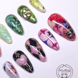 May include: A collection of artificial nails with various designs. The nails feature a range of colors, including green, pink, black, and purple, with gold accents. Some nails have intricate patterns, while others display cartoon-style illustrations. The "Kutty Handmade Nails" logo is visible.