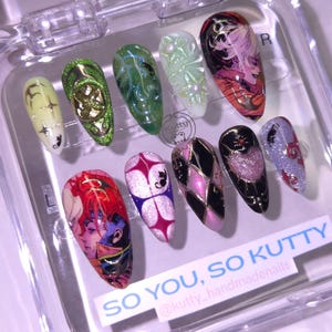 May include: A set of ten artificial nails in a clear plastic case. The nails feature various designs, including anime-style characters, geometric patterns, and glitter accents. Colors include pink, purple, black, gold, green, and white. Text on the case reads "SO YOU, SO KUTTY".