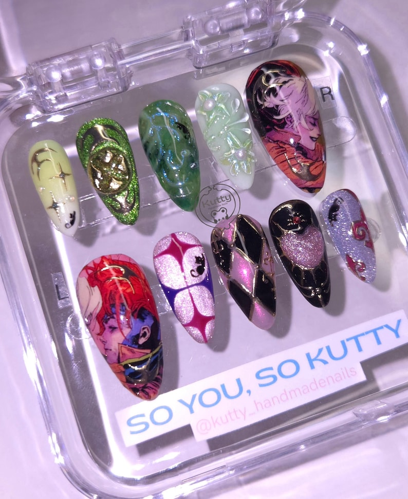 May include: A set of ten artificial nails in a clear plastic case. The nails feature various designs, including anime-style characters, geometric patterns, and glitter accents. Colors include pink, purple, black, gold, green, and white. Text on the case reads "SO YOU, SO KUTTY".