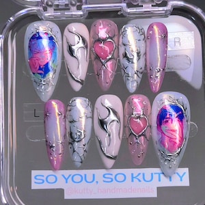 May include: A set of ten artificial nails in a clear case. The nails feature various designs, including pink hearts, silver accents, and a colorful anime-style image. The text "SO YOU, SO KUTTY" is visible.