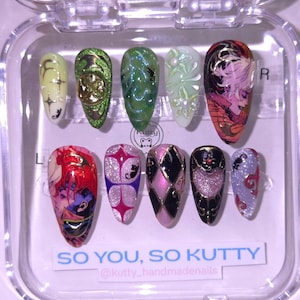 May include: A set of ten artificial nails in a clear plastic case. The nails feature various designs, including floral patterns, geometric shapes, and anime-style illustrations. Colors include green, pink, purple, black, and gold. Text on the case reads "SO YOU, SO KUTTY".