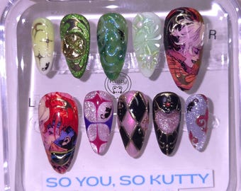 May include: A set of ten artificial nails in a clear plastic case. The nails feature various designs, including floral patterns, geometric shapes, and anime-style illustrations. Colors include green, pink, purple, black, and gold. Text on the case reads "SO YOU, SO KUTTY".