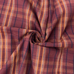 May include: Close-up of a burgundy fabric with a plaid pattern. The plaid design features vertical and horizontal lines in orange, navy blue, and white. The fabric is folded, creating a textured appearance.