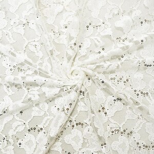 May include: Close-up of white lace fabric with a floral pattern and scattered sequins. The fabric is gathered in a swirl, showcasing its texture and design. Ideal for apparel and craft projects.