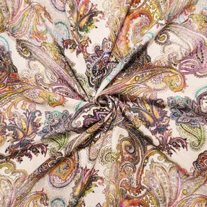 May include: A fabric featuring a dense, intricate paisley pattern in shades of purple, yellow, teal, orange, and black set against a light cream background. The material is gathered in the center, creating folds that highlight the detailed print.