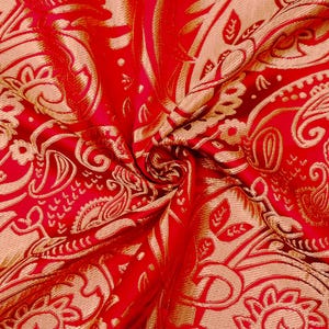May include: Close-up of a vibrant red fabric with an intricate paisley pattern in gold. The fabric is folded, creating a textured surface with swirling patterns and floral motifs. The design is detailed and ornate.
