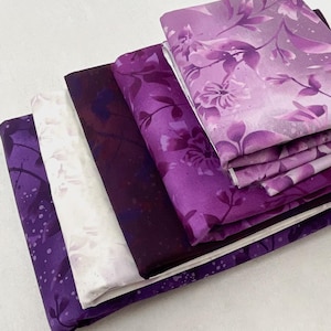 May include: A stack of fabric pieces in various shades of purple, ranging from deep violet to light lavender. The fabrics feature floral and abstract patterns, with some displaying speckled designs. The arrangement showcases the different textures and color variations.