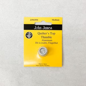 May include: A medium-sized John James Quilter's Top Thimble made of aluminum. The thimble is on a yellow card with the product name and brand. The card also includes the text "Made in Spain" and "Finest Quality Thimbles."