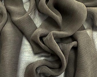 May include: Close-up of a flowing fabric with a textured, pleated appearance. The fabric is a muted olive green color, with areas of lighter gray tones. The material is draped and folded, creating shadows and highlights.