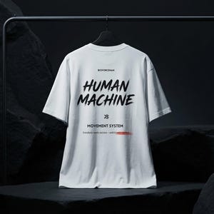 K&ouml;nnte beinhalten: Wei&szlig;es T-Shirt mit den Worten "HUMAN MACHINE" in gro&szlig;en, schwarzen Buchstaben im Pinselstil. Darunter der Text "MOVEMENT SYSTEM" und eine rote Linie. Das Shirt h&auml;ngt an einem schwarzen Kleiderb&uuml;gel vor dunklem Hintergrund.