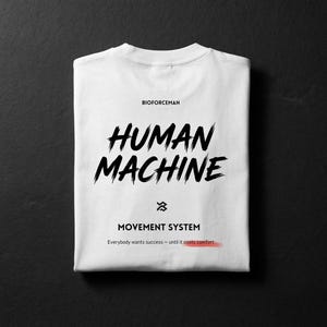 K&ouml;nnte beinhalten: Gefaltetes wei&szlig;es T-Shirt mit dem Schriftzug "HUMAN MACHINE" in schwarzer Pinselstrich-Schrift. Darunter stehen die Worte "MOVEMENT SYSTEM" und der Satz "Everybody wants success - until it costs comfort." Das T-Shirt liegt auf schwarzem Hintergrund.