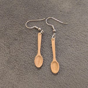 May include: A pair of earrings featuring miniature wooden spoon charms. The spoons are light brown with a small bowl and a long handle, suspended from silver-colored ear wires. The earrings are displayed on a gray, textured surface.