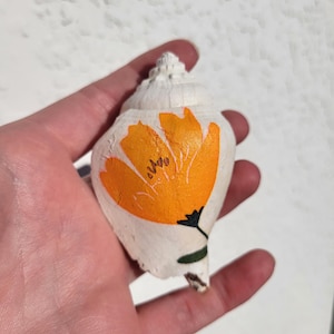 May include: A white seashell with an orange flower design. The flower has black details and a green stem. The seashell is held in a hand against a white background. The seashell is a decorative item.