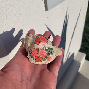 Puede incluir: Una concha beige decorada con un diseño floral con rosas rojas y rosas y hojas verdes. La concha se sostiene en una mano, mostrando los intrincados detalles de la obra de arte.