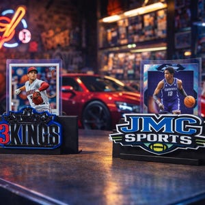 Puede incluir: Dos expositores de tarjetas deportivas con tarjetas de béisbol y baloncesto. Los expositores muestran los logotipos "3 Kings" y "JMC Sports". Un coche deportivo rojo es visible en el fondo. Las tarjetas están en fundas protectoras.