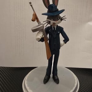 Può includere: Una statuetta di un personaggio coniglio che indossa un abito blu scuro, un fedora abbinato e una camicia bianca con cravatta. Il coniglio ha un sigaro in bocca e tiene in mano un mitra argentato e marrone su una base circolare.