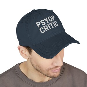May include: A navy blue distressed baseball cap with the white text "PSYOP CRITIC" across the front. The cap has a curved brim and a worn, vintage look.