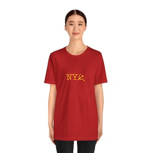Peut inclure: T-shirt rouge avec l'inscription dorée "NYC" et un symbole de la faucille et du marteau. Le t-shirt est à manches courtes et col rond.