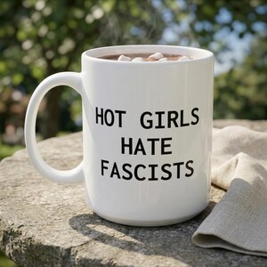 May include: A white ceramic mug with a handle, featuring the black text "HOT GIRLS HATE FASCISTS" printed on the front. The mug is filled with a brown liquid and several white marshmallows floating on the surface.