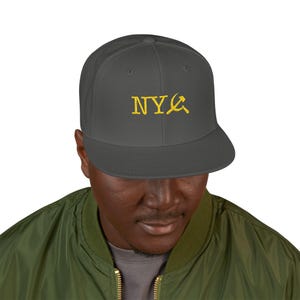 Puede incluir: Gorra de béisbol gris de ala plana con las letras "NY" y un símbolo de martillo y hoz dorado. La gorra la lleva una persona que lleva una chaqueta bomber verde.