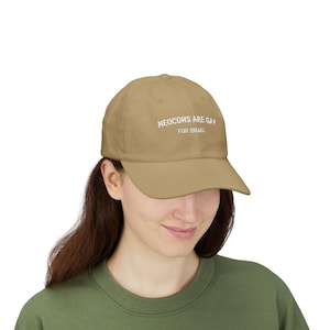 May include: A tan baseball cap with the text "NEOCONS ARE GAY FOR ISRAEL" in white. The cap has a curved brim and a classic six-panel design. The hat is worn by a person wearing a green shirt.