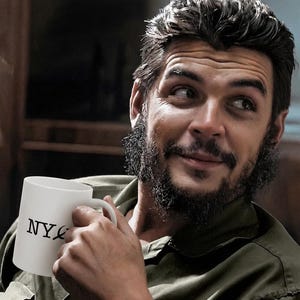 May include: A white ceramic mug with "NYC" printed in black, held by a person wearing an olive green shirt. The person has dark hair and a beard, looking off to the side. The image has a warm, inviting tone.