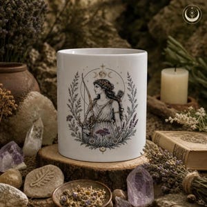 May include: A white ceramic mug featuring a detailed black and gold illustration of a woman with a bow and arrow, surrounded by a wreath of flowers and foliage, with a moon phase symbol above her head and a small deer standing beside her.