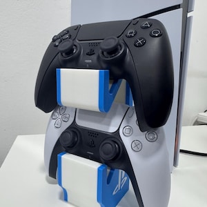 以下が含まれることがあります： 白と黒のPlayStation 5 DualSenseコントローラーが、白と青のコントローラー用スタンドに置かれています。スタンドにはPlayStationのロゴがあります。コントローラーは積み重ねられています。背景にはコンソールが見えます。