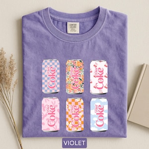 May include: Violet-colored t-shirt featuring six Diet Coke can designs. The cans showcase various patterns, including checkered, floral, and cloud motifs, in pink, orange, and blue hues. The shirt has a Comfort Colors tag.