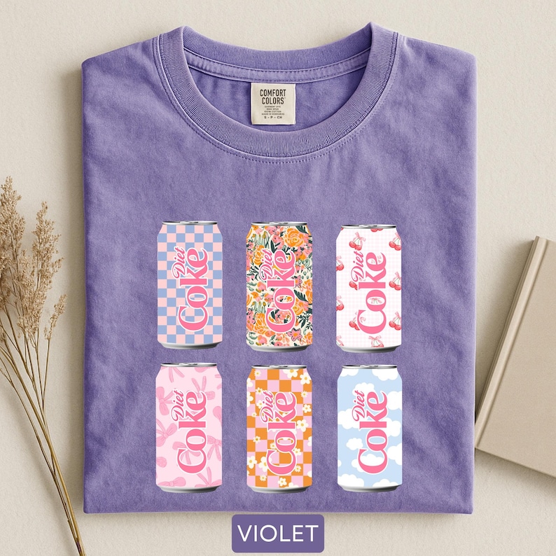 May include: Violet-colored t-shirt featuring six Diet Coke can designs. The cans showcase various patterns, including checkered, floral, and cloud motifs, in pink, orange, and blue hues. The shirt has a Comfort Colors tag.