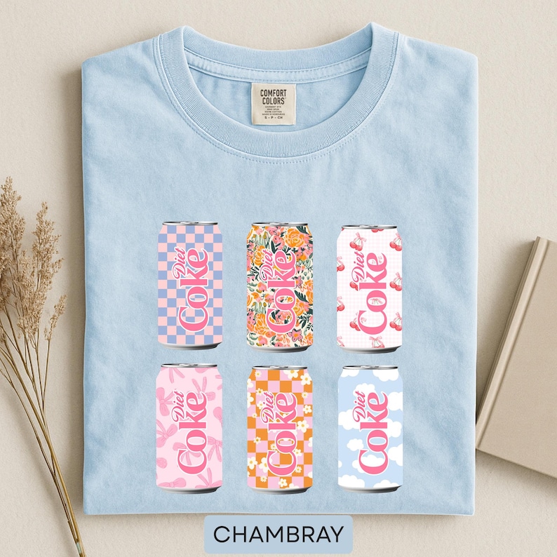 May include: Light blue t-shirt with six Diet Coke can designs. The cans feature various patterns, including floral, checkered, and cloud motifs, in pink, orange, and blue. The word "CHAMBRAY" is printed at the bottom.