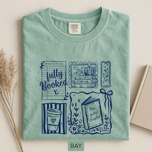 May include: A pale green t-shirt with a navy blue graphic design. The design includes a library book card, coffee cup, candle, and an open book with the words "One more chapter." The shirt also has the words "fully booked."