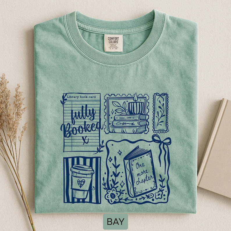 May include: A pale green t-shirt with a navy blue graphic design. The design includes a library book card, coffee cup, candle, and an open book with the words "One more chapter." The shirt also has the words "fully booked."