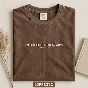 May include: Espresso-colored t-shirt with the text "FATHERHOOD IS KINGDOM WORK" and "PROVERBS 22:6" in white. The shirt has a crew neck and a tag that reads "COMFORT COLORS". The shirt is folded neatly on a light-colored surface.