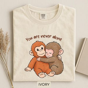 May include: Ivory-colored t-shirt featuring a cartoon illustration of two hugging monkeys. The text "You are never alone" is printed above the monkeys. The shirt has a crew neck and short sleeves.