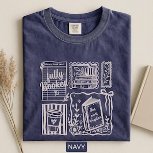 May include: Navy blue t-shirt with a white graphic design featuring a library book card, coffee cup, and an open book with the words "One more chapter." The shirt also has the words "fully booked."