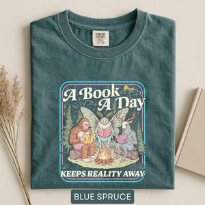 May include: A blue spruce t-shirt featuring a graphic of Bigfoot, a moth, and a deer-like creature sitting around a campfire reading books. The text reads A BOOK A DAY KEEPS REALITY AWAY. The design includes pine trees, stars, and a crescent moon.