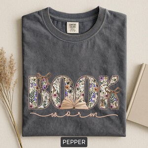 May include: A dark gray t-shirt with the word "BOOK" in a floral design, an open book graphic, and the word "worm" in a cursive font. The shirt is labeled "Pepper" at the bottom.