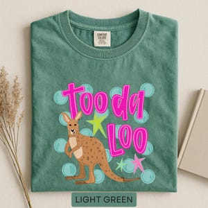 Op de afbeelding: Lichtgroen T-shirt met een kangoeroe-afbeelding en de tekst "Tooda Loo" in roze. De kangoeroe is bruin met een lichtbruine buik. Het ontwerp bevat blauwe bellen en steraccenten. De tekst "LIGHT GREEN" staat onderaan.