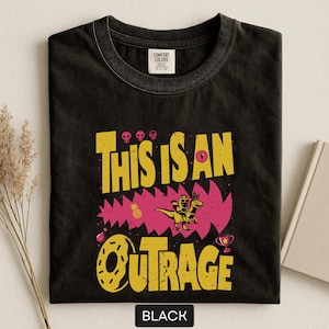 May include: Black t-shirt with a distressed graphic design. The design features the text "THIS IS AN OUTRAGE" in yellow, with a pink jagged background and a cartoon dinosaur riding another dinosaur. The word "BLACK" is at the bottom.