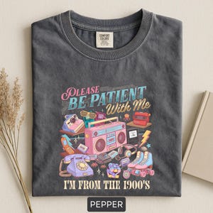 May include: Pepper-colored t-shirt with a retro graphic design. The design includes a pink boombox, roller skates, a rotary phone, and the text "PLEASE BE PATIENT WITH ME I'M FROM THE 1900'S."