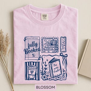 May include: A pale pink t-shirt with a navy blue graphic featuring a coffee cup, open book, and a stack of books. The text "fully booked" and "One more chapter" are included. The word "Blossom" is at the bottom.