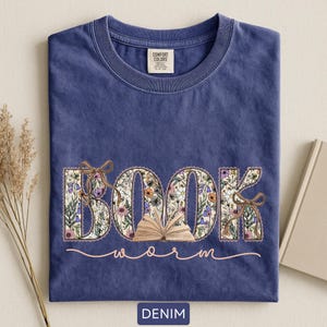 May include: A denim blue t-shirt with the word "BOOK" in floral print, an open book graphic, and the word "worm" in a cursive font. The shirt has a crew neck and a tag that says "Comfort Colors".